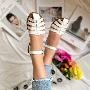 Puede incluir: Sandalias blancas de cuero con un dise&ntilde;o enjaulado y correas para el tobillo. Las sandalias las lleva una persona con las piernas desnudas y pantalones vaqueros azules.