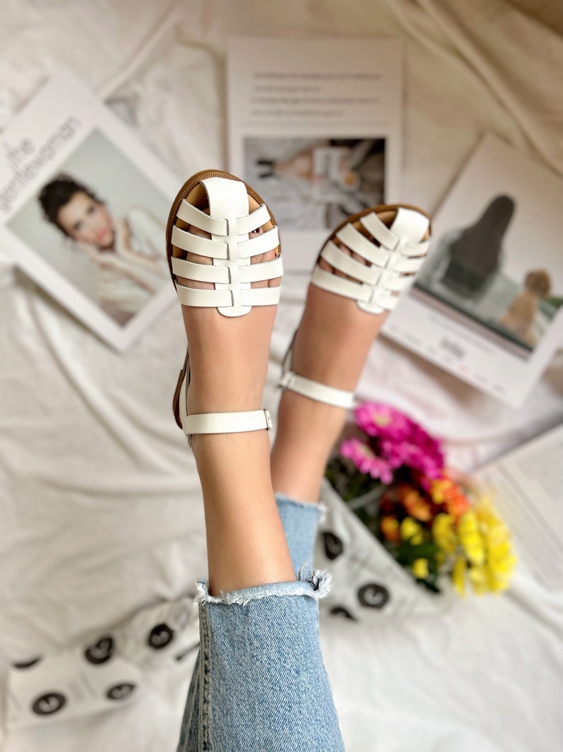 Puede incluir: Sandalias blancas de cuero con un dise&ntilde;o enjaulado y correas para el tobillo. Las sandalias las lleva una persona con las piernas desnudas y pantalones vaqueros azules.