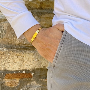 May include: A gold-toned metal bracelet with a flat, smooth surface. The bracelet is worn on the wrist of a person wearing a white shirt and gray pants.