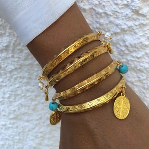 Pode incluir: Quatro pulseiras de ouro com um design texturizado, cada uma com um amuleto pendurado. Dois amuletos s&atilde;o contas azuis turquesa, um &eacute; uma p&eacute;rola branca e outro &eacute; um amuleto de ouro com uma cruz.