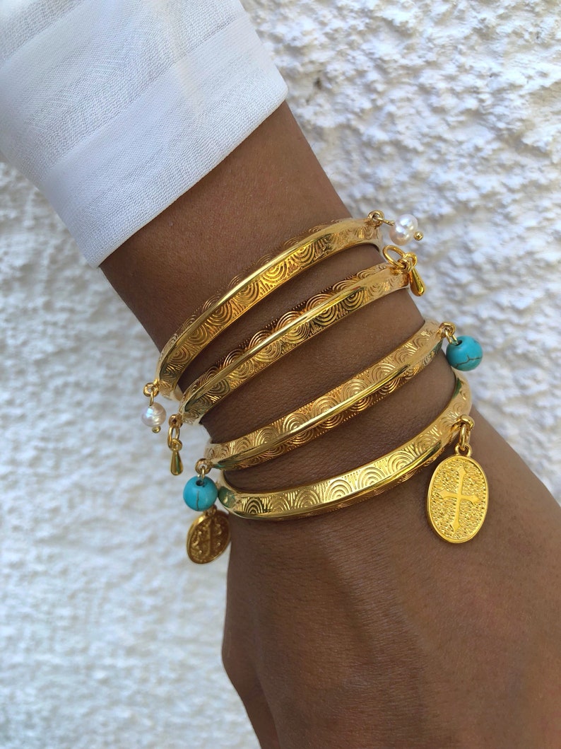 Pode incluir: Quatro pulseiras de ouro com um design texturizado, cada uma com um amuleto pendurado. Dois amuletos s&atilde;o contas azuis turquesa, um &eacute; uma p&eacute;rola branca e outro &eacute; um amuleto de ouro com uma cruz.
