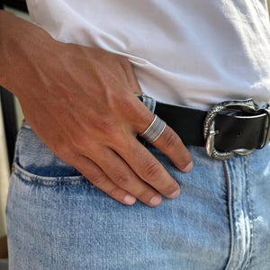 May include: A silver ring with multiple horizontal bands is worn on a hand. The person is wearing a white t-shirt, blue jeans, and a black belt with a decorative silver buckle.