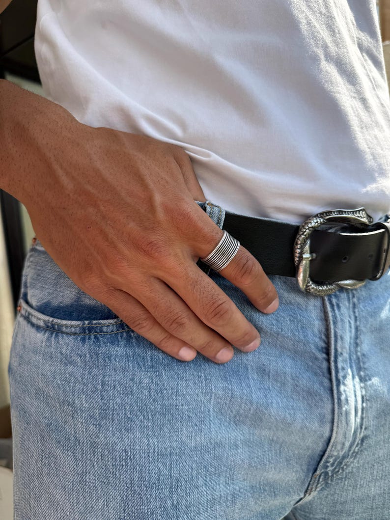 May include: A silver ring with multiple horizontal bands is worn on a hand. The person is wearing a white t-shirt, blue jeans, and a black belt with a decorative silver buckle.