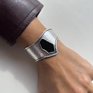 Op de afbeelding: Zilveren manchetarmband met een gehamerde textuur en een zwarte, geometrische middensteen. De armband is breed en wikkelt zich om de pols, wat een gedurfd, modern ontwerp laat zien. De manchet wordt om de pols van een persoon gedragen.