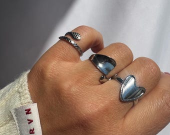 Puede incluir: Una mano muestra tres anillos plateados. Uno tiene un diseño de serpiente, otro una forma curva y lisa, y el tercero una forma de corazón. Los anillos están en una mano que lleva un suéter color crema con una etiqueta "KRVN."