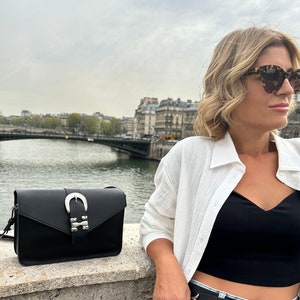 Puede incluir: Una mujer con una camisa blanca, un top negro corto y pantalones vaqueros azules con una hebilla de cintur&oacute;n plateada. Lleva un bolso de hombro de cuero negro con una hebilla plateada. El bolso tiene un dise&ntilde;o en forma de herradura.
