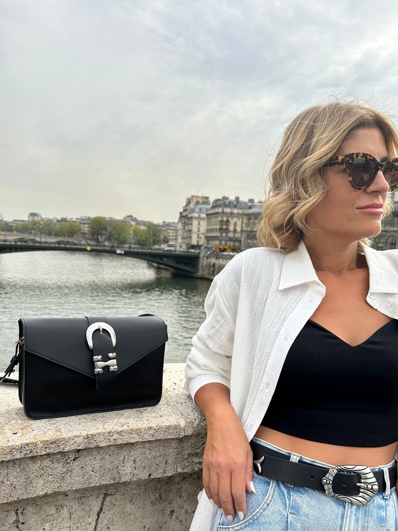 Puede incluir: Una mujer con una camisa blanca, un top negro corto y pantalones vaqueros azules con una hebilla de cintur&oacute;n plateada. Lleva un bolso de hombro de cuero negro con una hebilla plateada. El bolso tiene un dise&ntilde;o en forma de herradura.