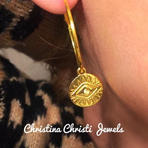 Puede incluir: Pendiente de aro dorado con un dije circular con un dise&ntilde;o de ojo. El dije tiene un borde texturizado y una peque&ntilde;a gema. El pendiente se muestra en una oreja. El texto "Christina Christi Jewels" es visible.