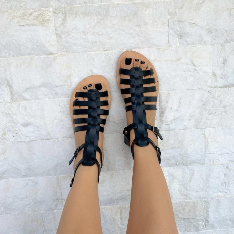 May include: A pair of black gladiator sandals featuring multiple horizontal straps across the foot, a vertical center strap, and an adjustable ankle strap with a metal buckle. The sandals have a flat sole and an open-toe design with black nail polish.