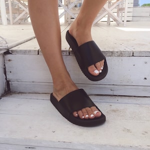 May include: A pair of black leather slide sandals with a thick sole. The sandals are worn on a person's feet.