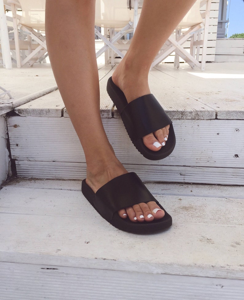 May include: A pair of black leather slide sandals with a thick sole. The sandals are worn on a person's feet.