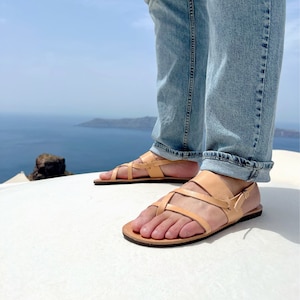 May include: A pair of tan leather sandals with a criss-cross strap design. The sandals are worn by a person standing on a white surface with a view of the ocean in the background.