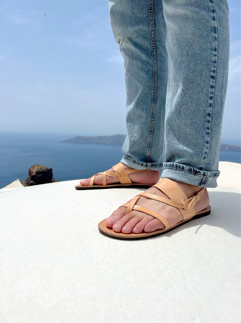 May include: A pair of tan leather sandals with a criss-cross strap design. The sandals are worn by a person standing on a white surface with a view of the ocean in the background.