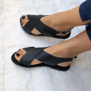 Puede incluir: Sandalias de cuero negro con tiras cruzadas y una tira para el tobillo. Las sandalias tienen una suela plana.
