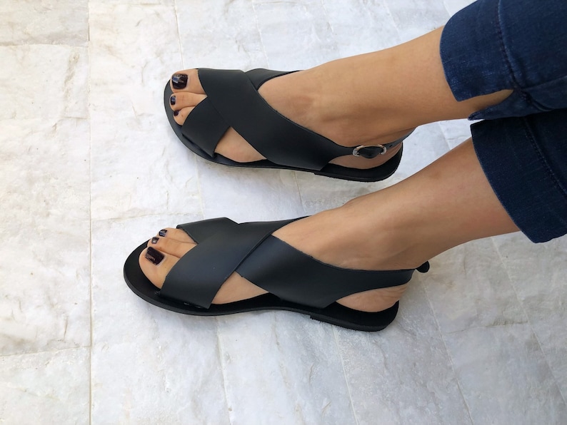 Puede incluir: Sandalias de cuero negro con tiras cruzadas y una tira para el tobillo. Las sandalias tienen una suela plana.