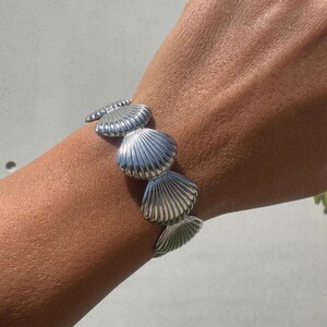 May include: A silver bracelet with seashell charms. The bracelet is worn on a wrist, showing the detailed texture and design of each shell. The shells are a bright silver colour. The bracelet is a piece of jewellery.