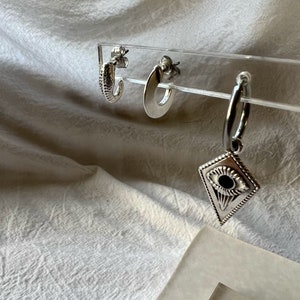 May include: Three silver hoop earrings. The earrings are displayed on a clear acrylic stand. The hoop earrings are all different sizes and styles. One earring has a small, round, eye-shaped charm.