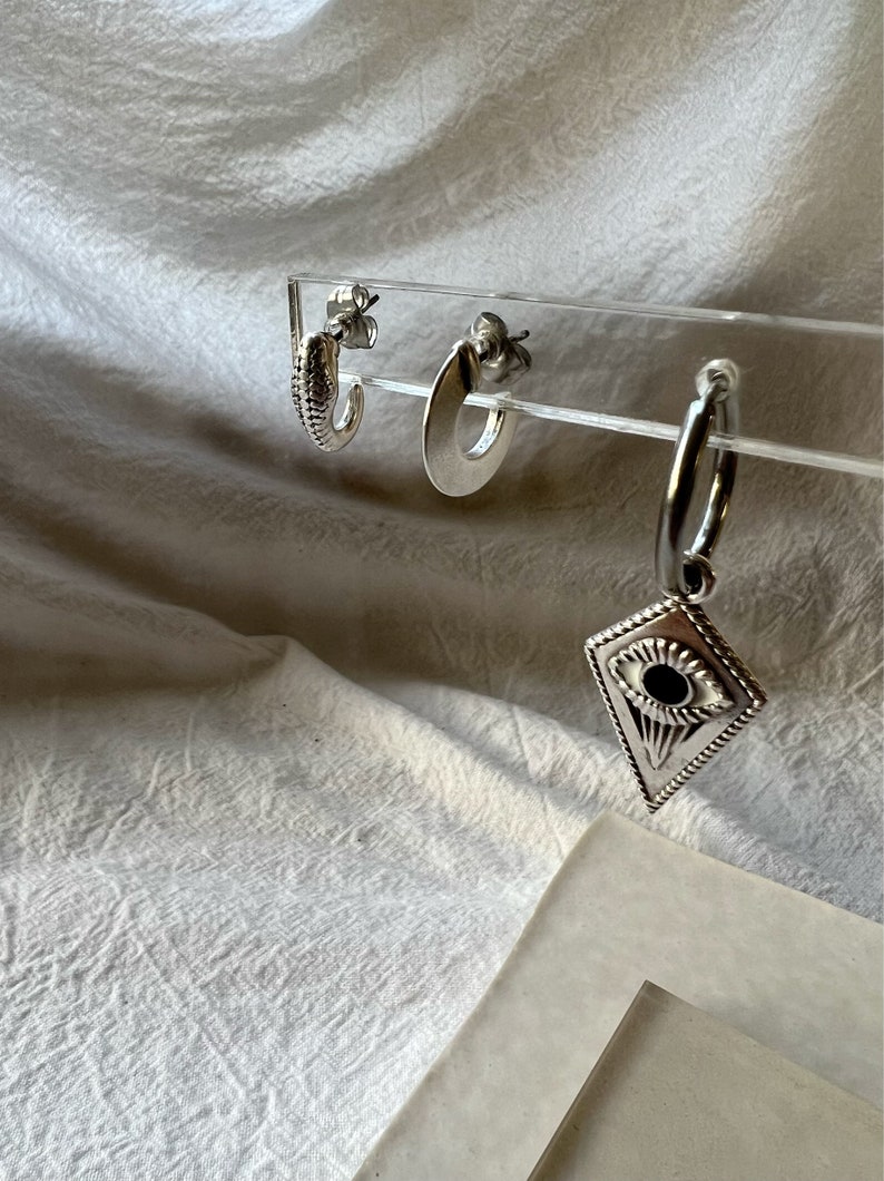 May include: Three silver hoop earrings. The earrings are displayed on a clear acrylic stand. The hoop earrings are all different sizes and styles. One earring has a small, round, eye-shaped charm.