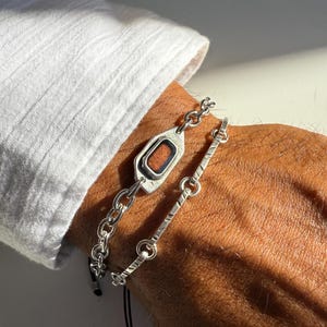 Op de afbeelding: Twee zilveren armbanden. Eén armband heeft een rechthoekige, roodbruine steen in een zilveren frame, verbonden door een ketting. De andere heeft een lineair ontwerp met een reeks kleine, verbonden zilveren staven. De armbanden worden om een pols gedragen, met een witte mouw op de achtergrond.