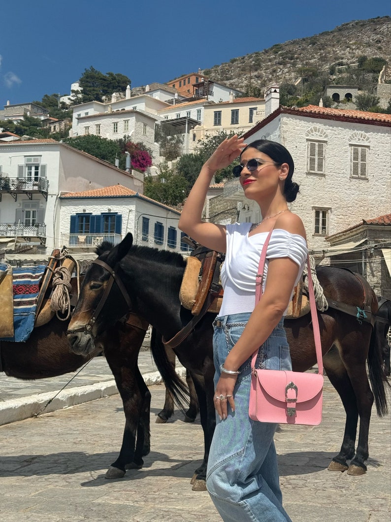 Op de afbeelding: Een vrouw in een wit, off-shoulder topje, blauwe jeans en zonnebril staat voor twee bruine ezels. Ze draagt een roze crossbody tas met een zilveren gesp. De ezels dragen zadels en staan op een geplaveide straat. De achtergrond is een wit gebouw met een witte muur.