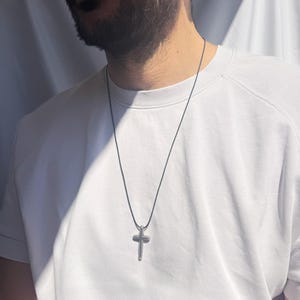 Peut inclure: Un pendentif en forme de croix argentée sur un collier en cordon noir. Le collier est porté par une personne avec une barbe.