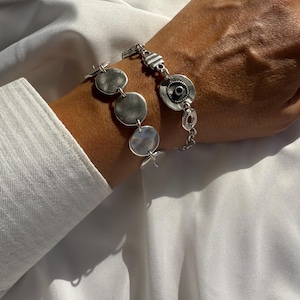 May include: A silver bracelet with a hammered disc design and a circular element with a black center. The bracelet is worn on a wrist, with a white shirt sleeve visible in the background. The bracelet is made of metal.