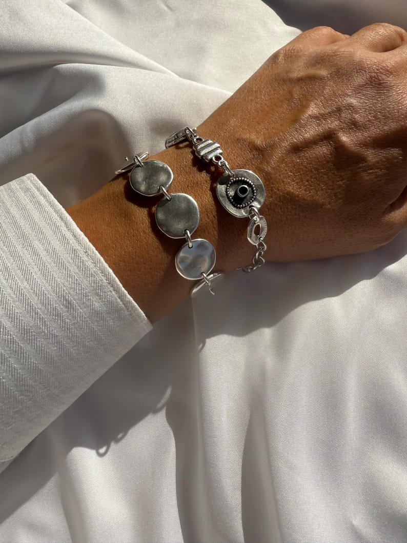 May include: A silver bracelet with a hammered disc design and a circular element with a black center. The bracelet is worn on a wrist, with a white shirt sleeve visible in the background. The bracelet is made of metal.