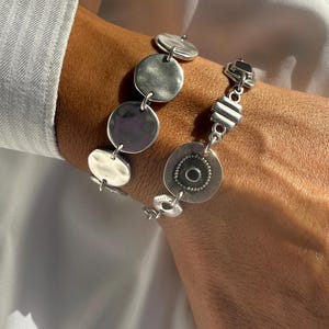 May include: Two silver bracelets are displayed on a wrist. One bracelet features large, flat, circular links. The other bracelet has a mix of shapes, including a large circle with a central design and smaller rectangular links. The bracelets are worn against a white shirt.