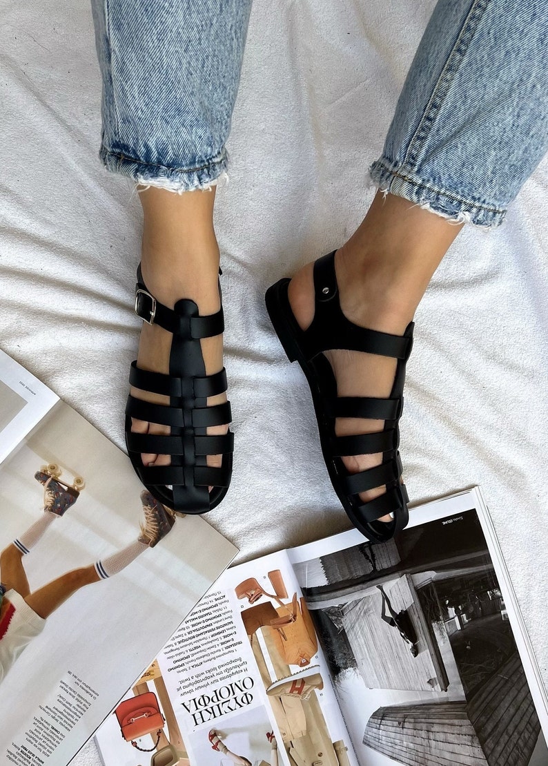 Op de afbeelding: Zwarte leren gladiatorsandalen met een gespsluiting. De sandalen hebben een kooiconstructie en een platte zool.