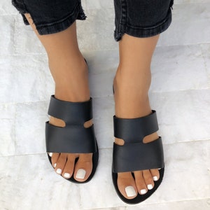Puede incluir: Sandalias de cuero negro con un dise&ntilde;o de doble correa. Las sandalias son de punta abierta y tienen una suela plana. La imagen muestra las sandalias desde una perspectiva superior, con los pies de una persona que las lleva.
