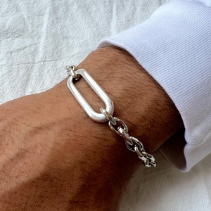 K&ouml;nnte beinhalten: Ein silbernes Kettenarmband mit einem gro&szlig;en ovalen Glied. Das Armband befindet sich an einem Handgelenk.