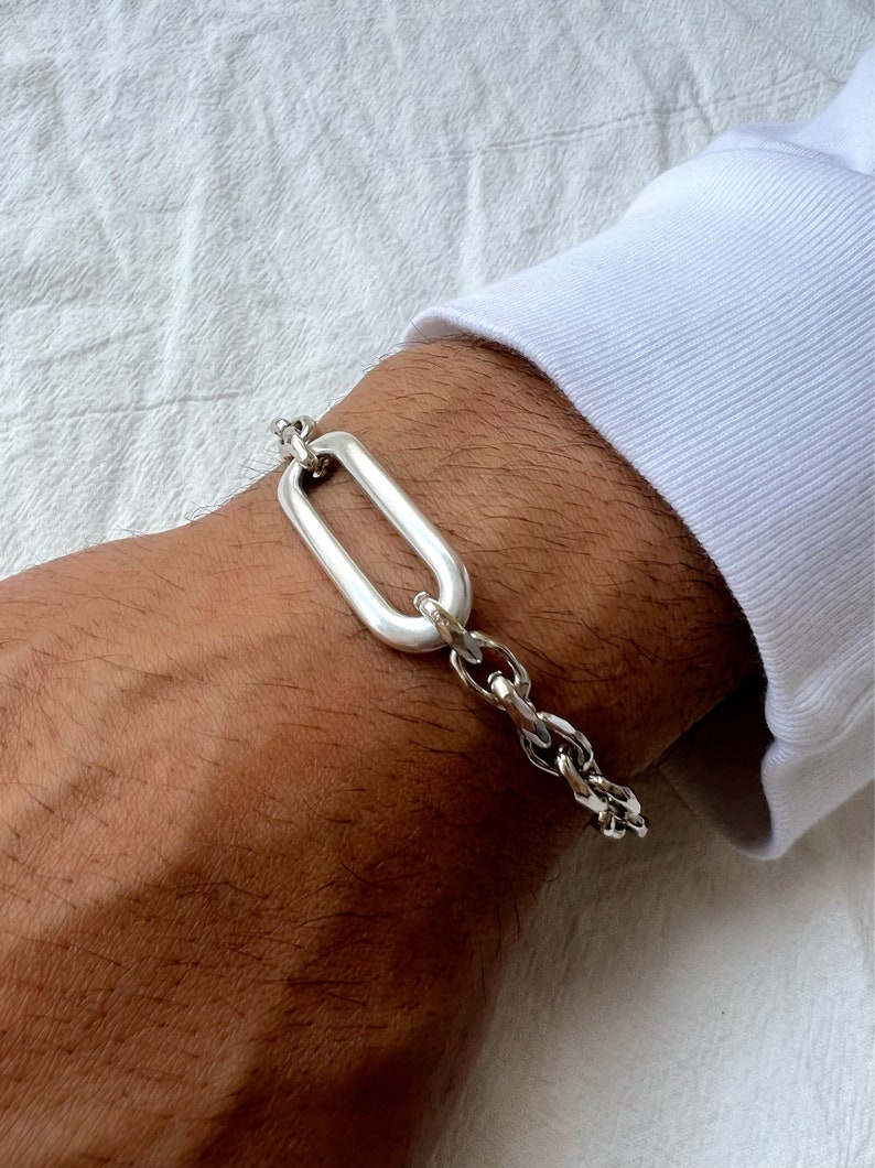 K&ouml;nnte beinhalten: Ein silbernes Kettenarmband mit einem gro&szlig;en ovalen Glied. Das Armband befindet sich an einem Handgelenk.