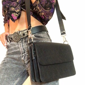 Puede incluir: Un bolso bandolera de cuero negro con un acabado texturizado y una hebilla plateada. El bolso tiene una correa larga ajustable y un cierre de solapa.