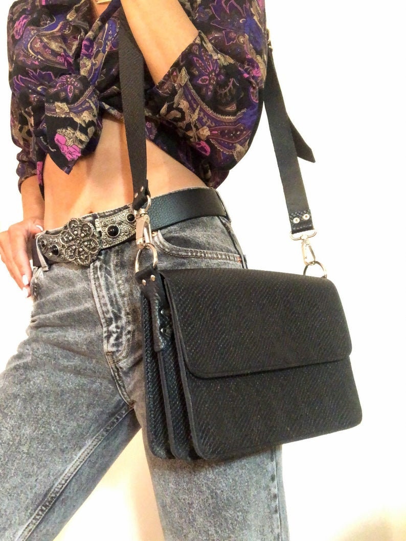 Puede incluir: Un bolso bandolera de cuero negro con un acabado texturizado y una hebilla plateada. El bolso tiene una correa larga ajustable y un cierre de solapa.