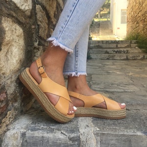 May include: Tan leather sandals with a cross-strap design and a woven platform sole. The sandals have a buckle closure and are worn with light wash denim jeans. The platform sole measures approximately 1.5 inches.