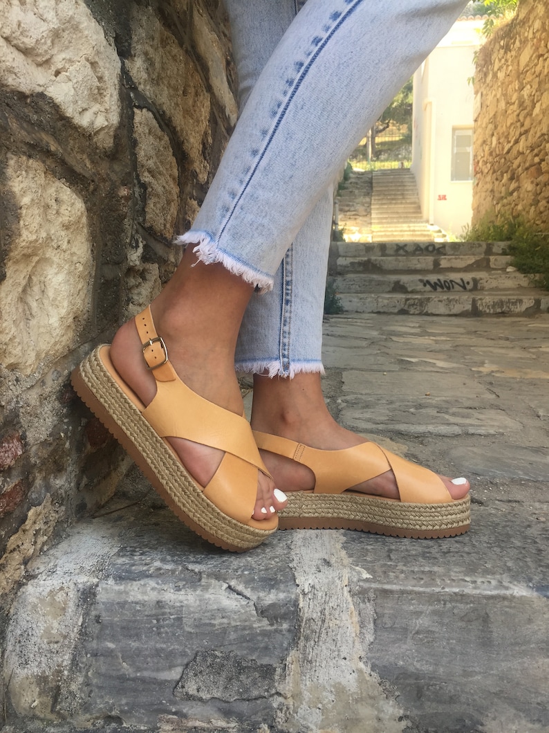 May include: Tan leather sandals with a cross-strap design and a woven platform sole. The sandals have a buckle closure and are worn with light wash denim jeans. The platform sole measures approximately 1.5 inches.