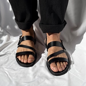 K&ouml;nnte beinhalten: Schwarze Ledersandalen mit mehreren Riemen und einem Schnallenverschluss. Die Sandalen haben eine runde Spitze und eine wei&szlig; gen&auml;hte Sohle. Die Person tr&auml;gt eine schwarze Hose mit hochgekrempelten Manschetten.