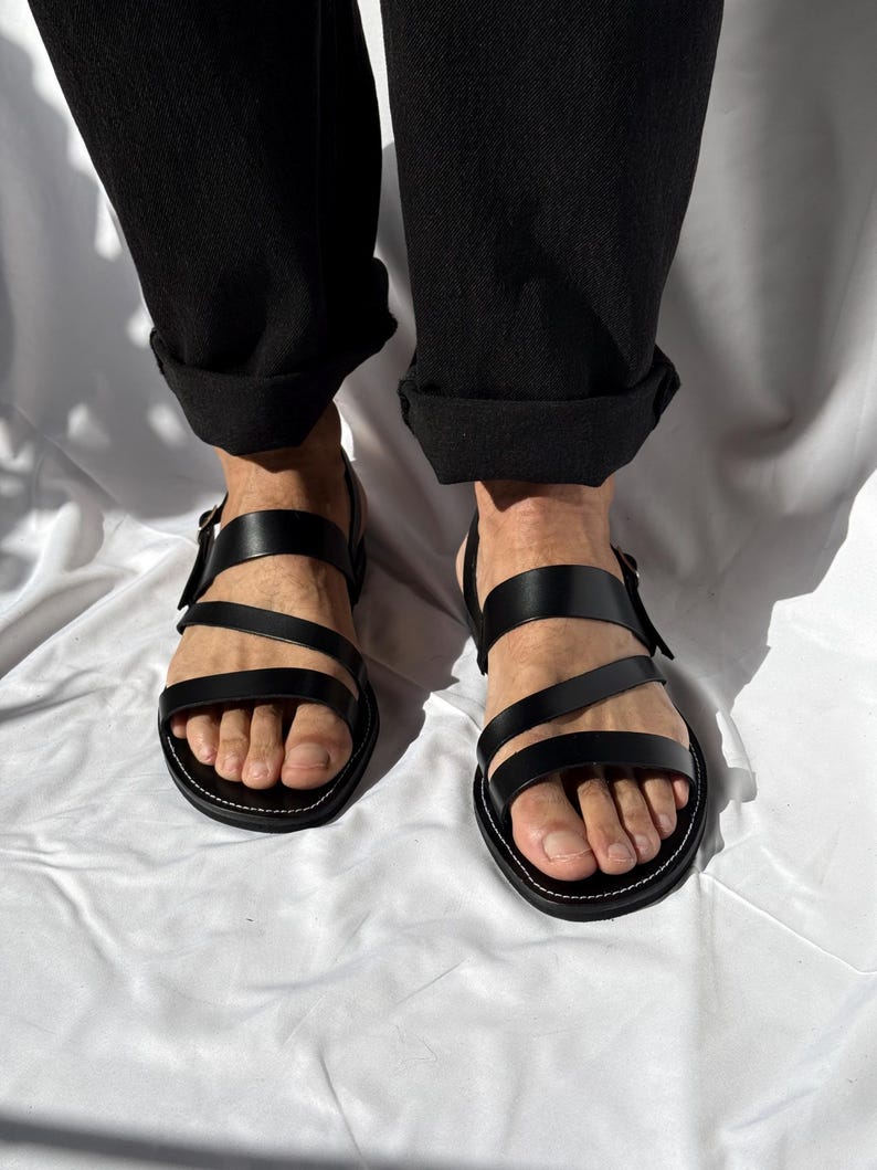 K&ouml;nnte beinhalten: Schwarze Ledersandalen mit mehreren Riemen und einem Schnallenverschluss. Die Sandalen haben eine runde Spitze und eine wei&szlig; gen&auml;hte Sohle. Die Person tr&auml;gt eine schwarze Hose mit hochgekrempelten Manschetten.