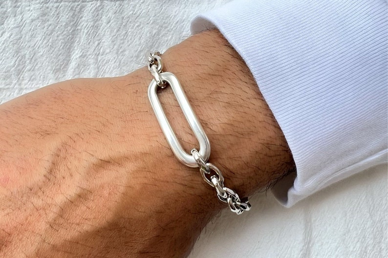 K&ouml;nnte beinhalten: Ein silbernes Kettenarmband mit einem gro&szlig;en ovalen Glied. Das Armband befindet sich an einem Handgelenk.