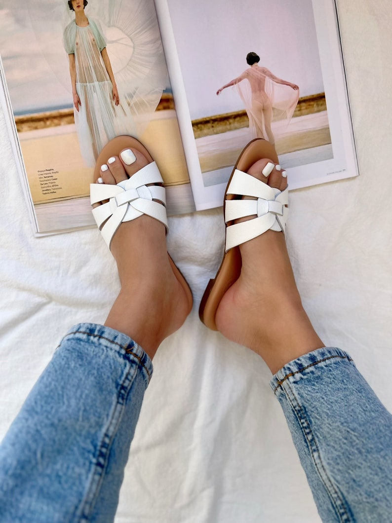 Puede incluir: Sandalias blancas de cuero con un dise&ntilde;o de correa cruzada. Las sandalias tienen una suela marr&oacute;n y las lleva puestas una persona. La persona lleva unos vaqueros azul claro.