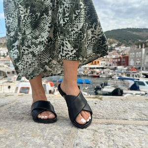 Puede incluir: Un par de sandalias de cuero negro con un dise&ntilde;o de correa cruzada. Las sandalias las lleva puesta una persona de pie sobre una superficie de piedra.