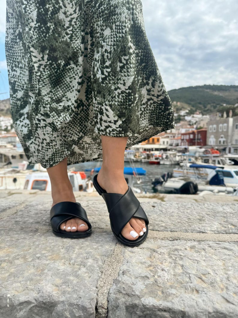 Puede incluir: Un par de sandalias de cuero negro con un dise&ntilde;o de correa cruzada. Las sandalias las lleva puesta una persona de pie sobre una superficie de piedra.