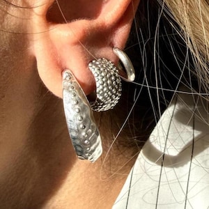 Può includere: Primo piano di tre orecchini d'argento. Uno è un cerchio grande e strutturato con un design martellato. Un altro è un cerchio più piccolo con una trama a pois. Il terzo è una piccola barra curva e liscia.