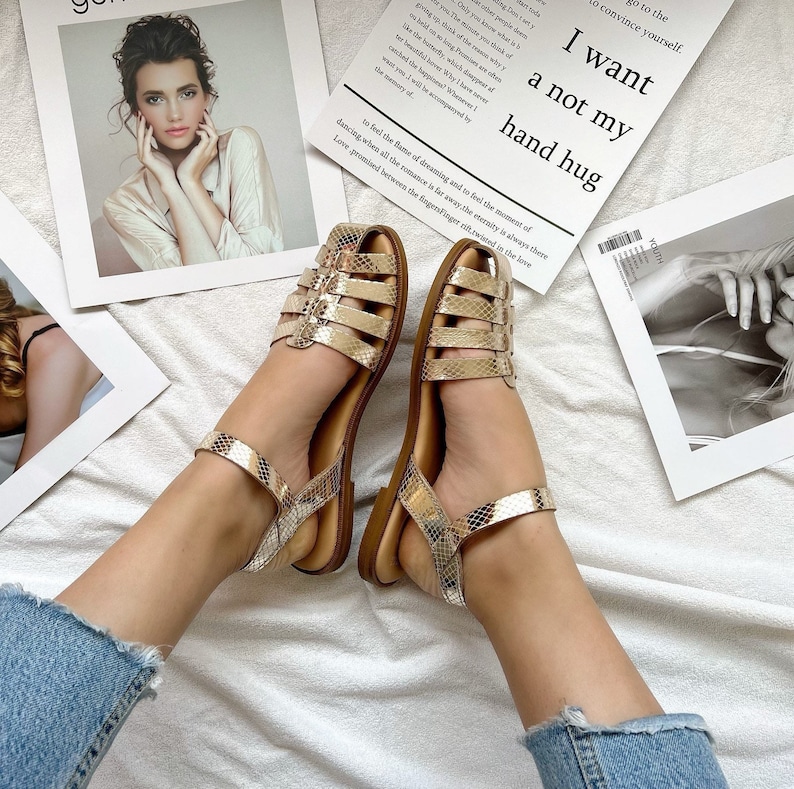 May include: A pair of gold sandals with a snake-skin pattern and a buckle closure. The sandals are on a white surface with a piece of paper that says "I want a not my hand hug".
