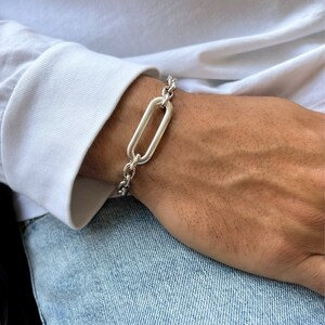 K&ouml;nnte beinhalten: Ein silbernes Kettenarmband mit einem gro&szlig;en ovalen Glied. Das Armband befindet sich an einem Handgelenk.