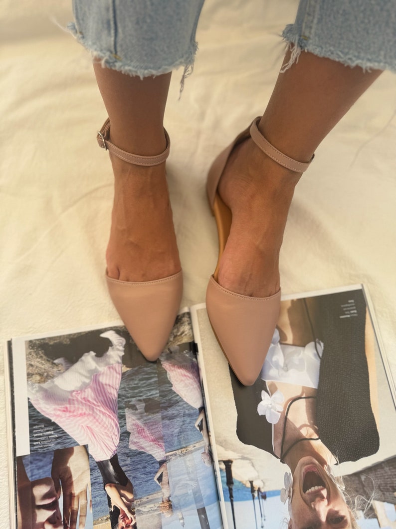 May include: A pair of blush-colored, pointed-toe flats with ankle straps. The shoes are displayed on top of a magazine with a collage of images. The flats have a simple, elegant design, suitable for various occasions.