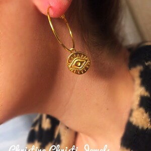 Puede incluir: Pendiente de aro dorado con un dije circular con un dise&ntilde;o de ojo. Tambi&eacute;n se ve un peque&ntilde;o pendiente de bot&oacute;n plateado en forma de V. El texto "Christina Christi Jewels" est&aacute; en la parte inferior de la imagen.