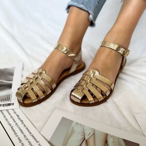 May include: Gold sandals with a snake-skin pattern and a buckle closure. The sandals have a flat sole and are made of leather.