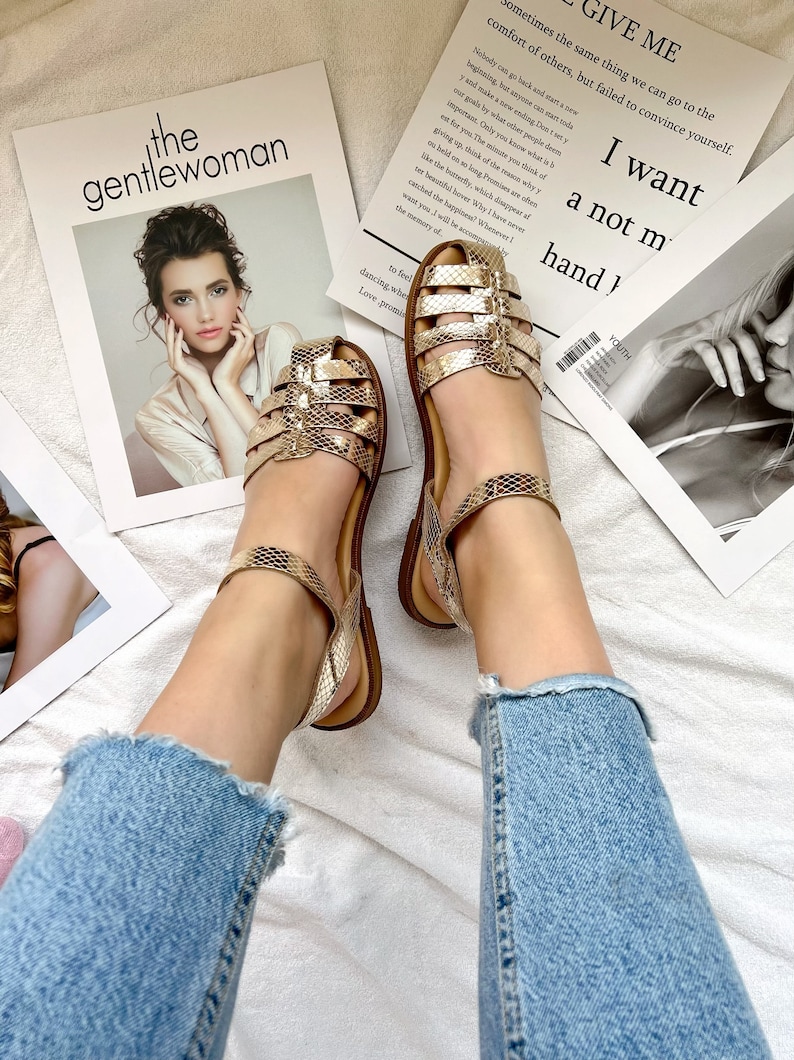 Puede incluir: Un par de sandalias doradas de piel de serpiente con una gruesa correa de tobillo. Las sandalias est&aacute;n sobre una superficie blanca con una revista que presenta el texto "the gentlewoman" y otra revista con el texto "GIVE ME sometimes the same thing we can go to the comfort of others, but failed to convince yourself. I want a not m hand)"