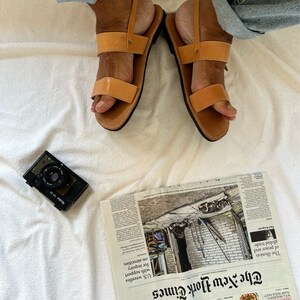 May include: A pair of brown leather sandals with two straps across the foot. The sandals are worn by a person wearing blue jeans. The sandals are on a white surface.